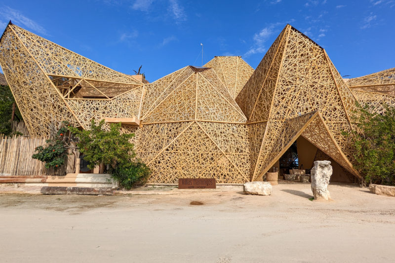 exterior of Hotel Villas Flamingo in Holbox, with unique geometric architecture featuring large pointed shapes built from very light coloured wood, in front of a sandy road