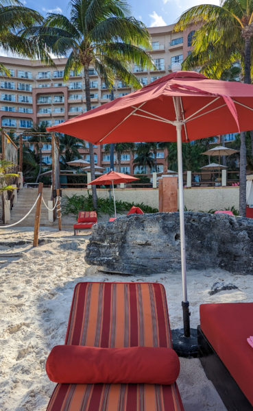 red and orange striped sun lounger under a red umbrella on a white sandy beach with palm trees behind and a large pink coloured hotel building behind that