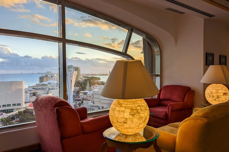 laerge amber coloured lamp with a glowing circular base on a table next to two red armchairs in front of a large window with a view of the Cancun Hotel Zone's skyscrapers and resorts outside and cloudy sunset sky overhead. 