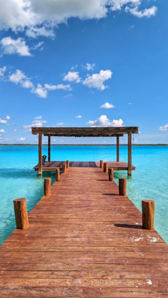 wooden jetty made from reddish coloured wood with a small covered structure at the end leading out into a bright blue lake with blue sky overhead. how to get to bacalar mexico.