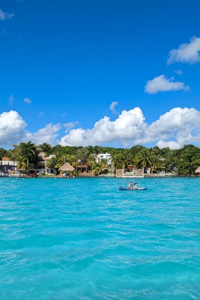 shot of a lake with vivie cyan water and the distant bank covered in palm trees with a few houses and buildings dotted between the trees. there are two people rowing a blue kayak on the lake i nthe distance