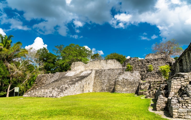 grey stone ruined pyramid with stone steps set in a grassy field wtih green leafy trees behind and a birght blue sky. Ruins of the ancient Mayan city of Kohunlich in Quintana Roo, Mexico