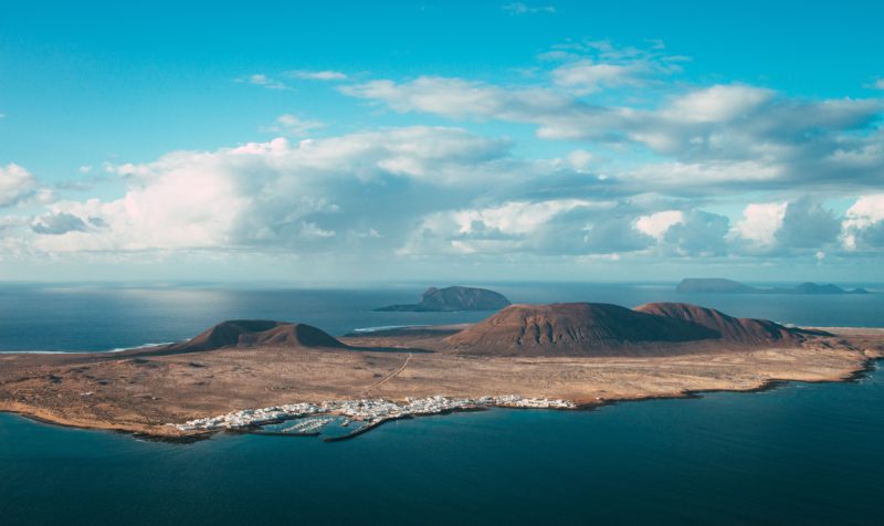 aerial shot of a small mainly brown/dusty island with some low brown mountains and a tiny town of white buildings on the edge closest, all surrounded by blue sea with bright blue sky and fluffy clouds above. where to stay in lanzarote.
