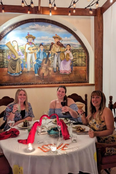 three girls sitting around a round table with white tablecloth lifting their glasses and smiling at the camera. there is a painting on the wall behind showign a traditional mexican band and large fairy lights hanging from the wooden beams overhead.