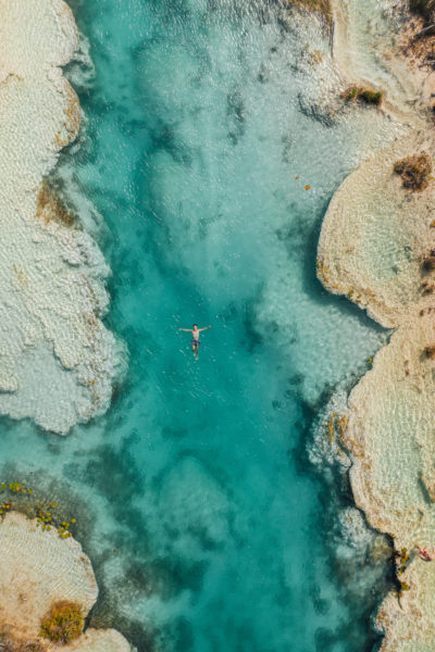 aerial shot of a narrow channel of turquoise water lined with grey and white rocky formations with a boy floating on top of the water