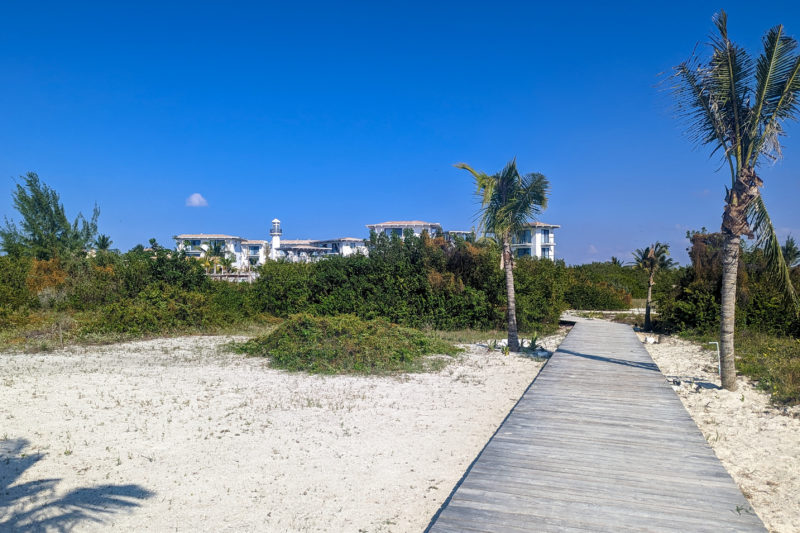 white sandy beach with some small bushes behind it and a long wooden walkway leading away between two palm trees, with a large white hotel building visible in the distance, on a very sunny day with clear blue sky above.