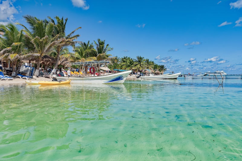 looking across very clear shallow sea water towards a narrow patch of white sandy beach with several small fishing boats near the shore and lots of palm trees behind on a very sunny day with blue sky above. Mahahual in mexico. 