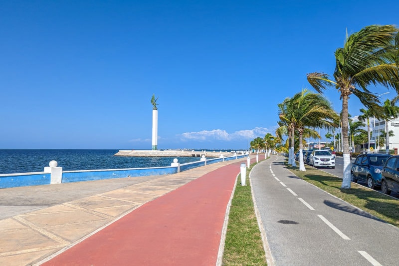 looking down an empty road next to a pink paved cycle path alongside a beige paved esplanade with palm trees lining the right side of the road and the sea on the left of the esplanade. in the