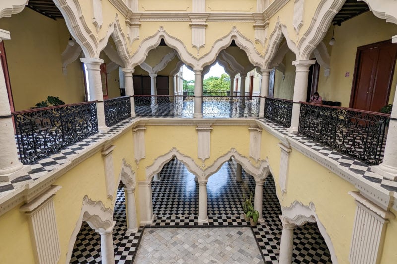 looking down from the 2nd floor at the central courtyard of Mansion Carvajal with a white marble floor surroudned by black and white diamond tiled floor. the walls are pale yellow with white archways all around both levels.