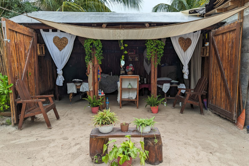 small one room massage parlour on the beach with sandy floor and open front and walls made of wood with three massage beds inside
