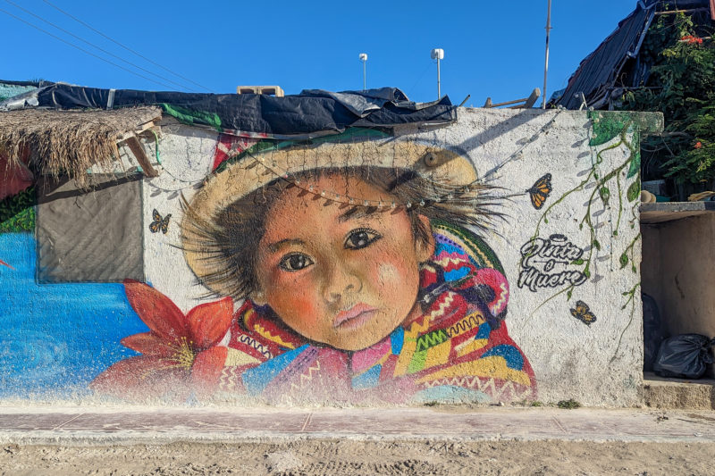 large mural on a white wall of a mexican child with a traditional colourful shawl and straw hat ona sunny day with blue sky above and a sandy road in front