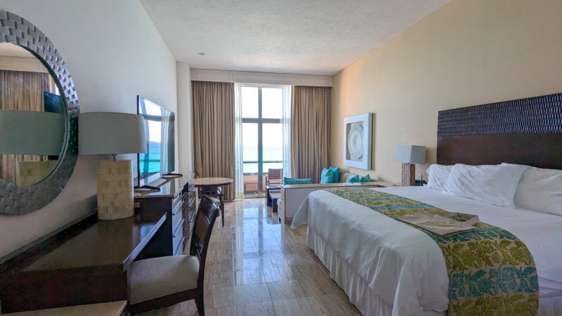 hotel bedroom with cream coloured marble floors and white walls and cieiling. there is a large white med with a yellow runner on it to the right and a large french door at the end of the room opening onto a balcony with a view of the ocean behind. Suite at the Grand Fiesta Americana Coral Beach Cancun - review