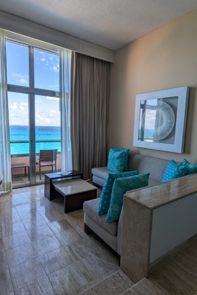 hotel room with cream coloured marble floor and a grey corner sofa with bright blue scatter cusions on it facing glass french doors to a balcony with vivid cyan sea visible below