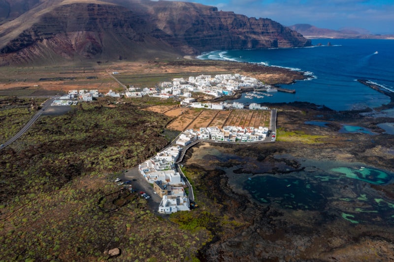 aerial shot of a small town full of white buildings on the coast with greenery on one side and the blue sea on the right and very tall, rugged dark brown cliffs behind