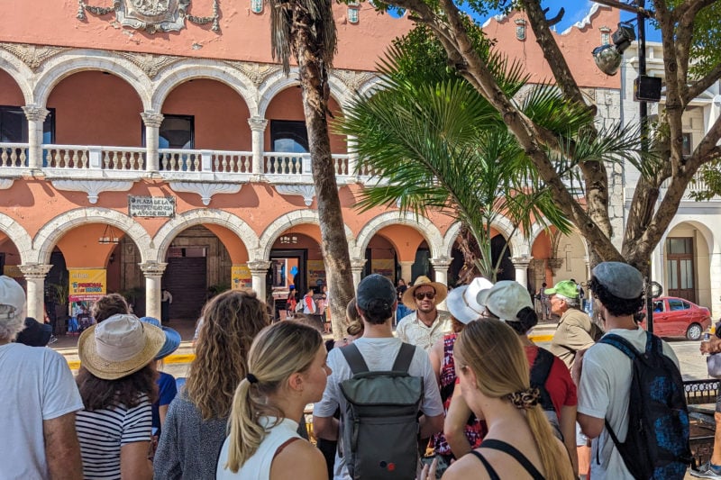 large group of people facing away from the camera towards a tour guide in front of a large peach coloured building with a row of arches on both the first and second floor