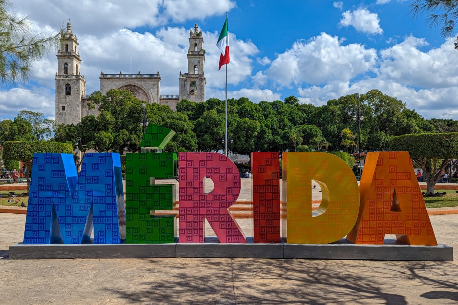 large sign with colourful 3D letters spelling out the name Mérida in a park with green bushes behind and the two towers of a white stone cathedral behind that on a bright sunny day with blue sky and fluffy clouds overhead. what to do in Mérida Mexico.