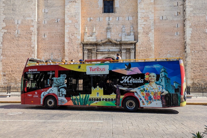 open top double decker bus with a huge multicoloured mural on the side showing different buildings from around Mérida and a woman in colourful traditional dress. the bus is parked in front of the Cathedral which has a carved stone frame aroudn the doorway and pinkish-cream coloured facade. 