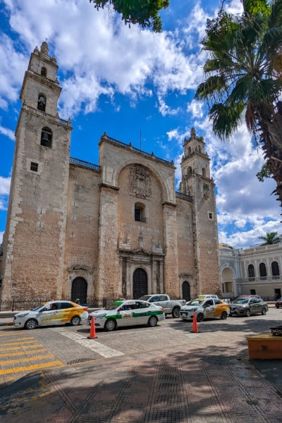 large cathedral built from light pink cream stone with two small towers and a large arch in the centre of the facade with a busy road full of queuing cars in front and the fronds of a palm tree in the top right corner. what to do in Mérida Mexico