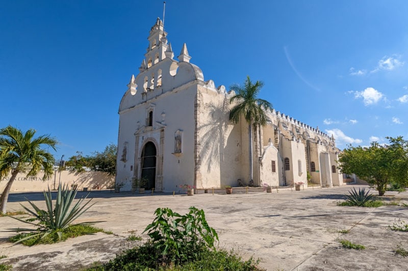small white church in a stone courtyard with several palm trees dotted around. what to do in merida mexico