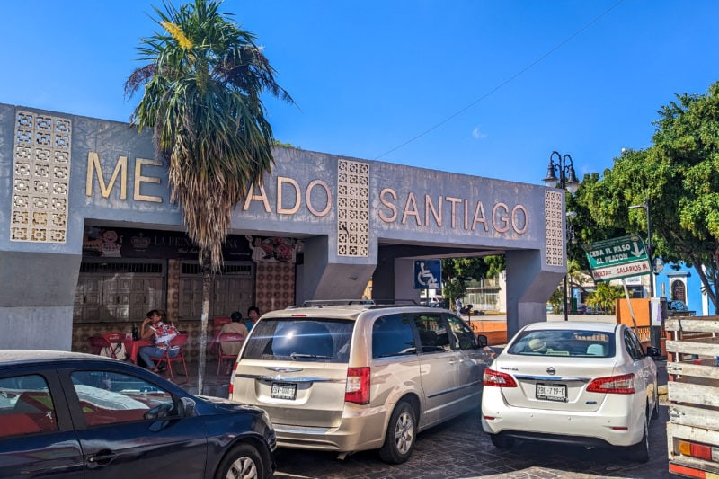 exterior of a one storey building with blue tiled walls and the words Mercado Santiago along the top in gold letters. there is a palm tree in front and several cars in the street in front of that and empty blue sky behind.