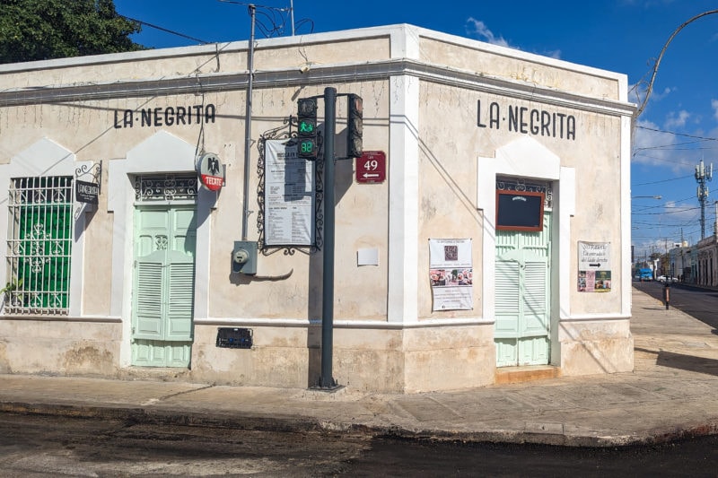 cream painted one storey building with light green saloon style swing doors and a small black sign over the doors that says La Negrita - a small cantina bar in Merida during the day 