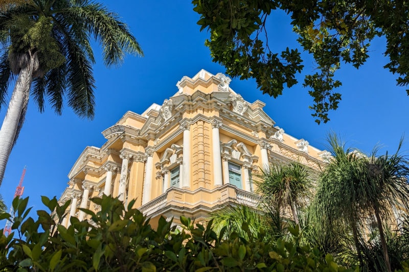 corner of a pastel-yellow palace with white trim and a carved facade framed by green bushes and trees against a clear blue sky