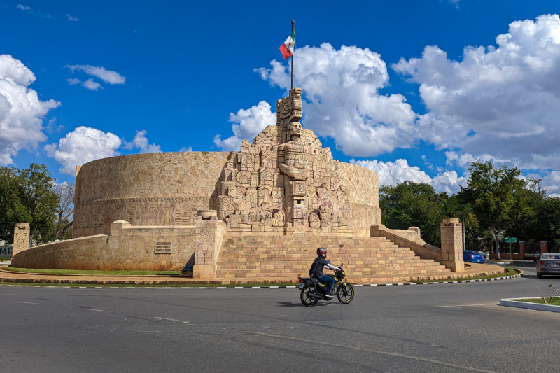 Large circular monument built from beige coloured stone with a Mayan statue carved on the front and the mexican flag flying from it. a motorbike is driving in front of the stone steps.