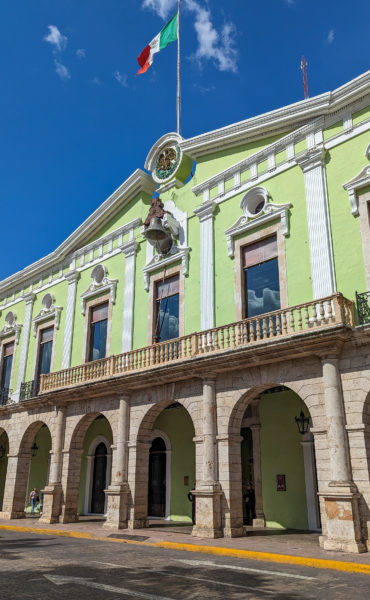 facade of a light green building with a triangular roof and  a row of stone arches along the ground floor