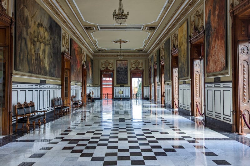 interior of a stately historic room in a palace with a black and white tiled floor and dark wood panelling along the walls with several large painted murals high up on the walls and two windows at the end. 