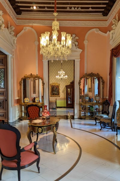 interior of an old colonial mansion with peach painted walls and a gold chandelier above two pink chairs made from dark wood with a white stone floor underneath - inside Casa Montejo Merida