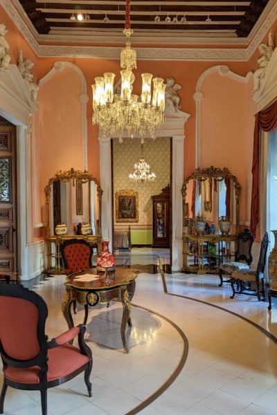 interior of an old colonial mansion with peach painted walls and a gold chandelier above two pink chairs made from dark wood with a white stone floor underneath - inside Casa Montejo Merida 