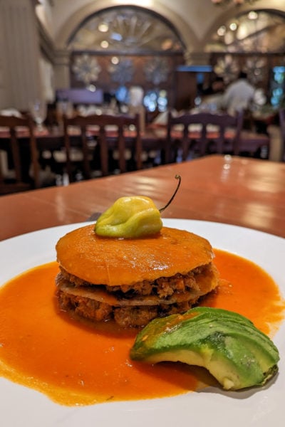 close up of a white plate with orange sauce on it and a small tower made of layers of three tortillas stuffed with minced fish meat and topped with a green habanero chilli with the restaurant out of focus in the background