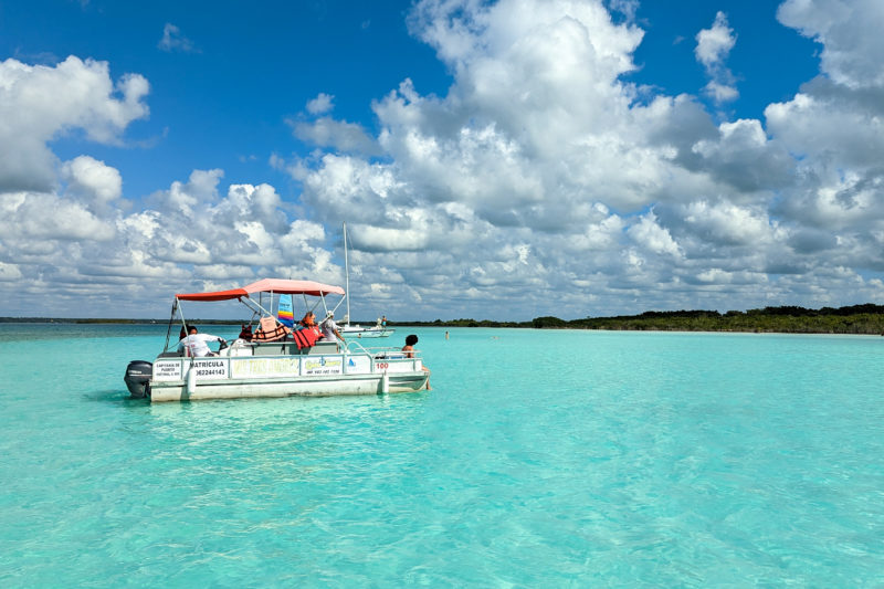 small white pontoon style boat with a red fabric roof floating on a lake with vivid cyan coloured water on a very sunny day with blue sky and fluffy white clouds overhead