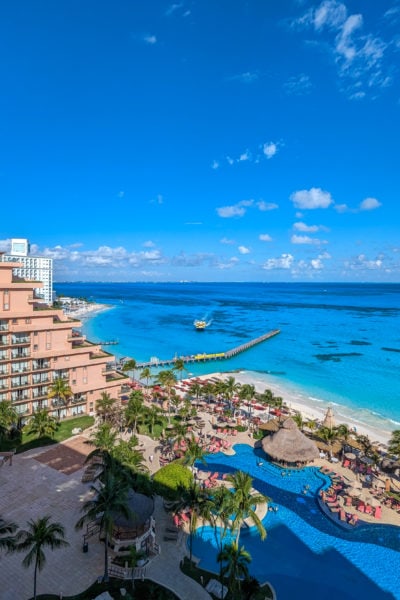 view from above of a large outdoor pool in frotn of a white sandy beach with the turquoise sea behind on a very sunny day. there is a narrow wooden pier to the left and on land in the left of the frame is the edge of a triangular shaped pinkish coloured 12 storey hotel. Grand Fiesta Americana Coral Beach Cancun review. 