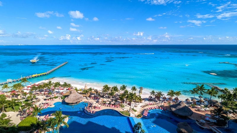 view from above of a large outdoor pool in frotn of a white sandy beach with the turquoise sea behind on a very sunny day. there is a narrow wooden pier to the left and a yellow board approaching. Grand Fiesta Americana Coral Beach Cancun review. 