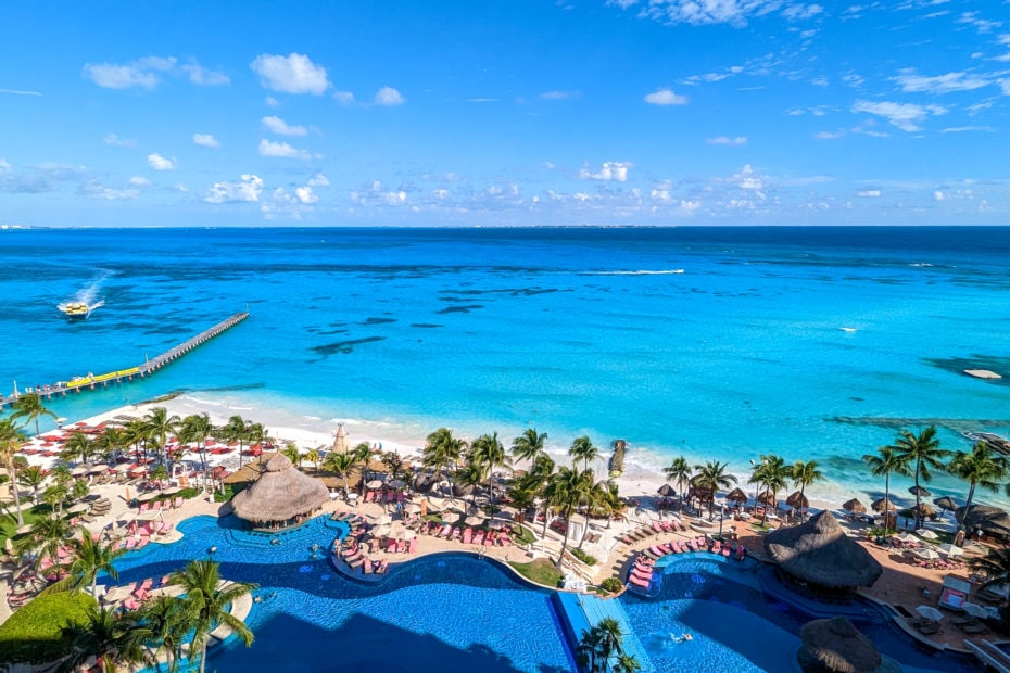 view from above of a large outdoor pool in frotn of a white sandy beach with the turquoise sea behind on a very sunny day. there is a narrow wooden pier to the left and a yellow board approaching. Grand Fiesta Americana Coral Beach Cancun review.