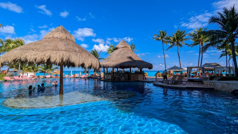 bright blue outdoor pool with a large grass roofed wooden umbrella at the centre and a grass roofed palapa style bar on the far side, with several palm trees to the right and a bright blue sky above. 