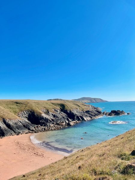 view of a golden sandy beach from a low grassy cliff with a small bay of turqouise water with low cliffs on the other side on a very sunny day with blue sky above