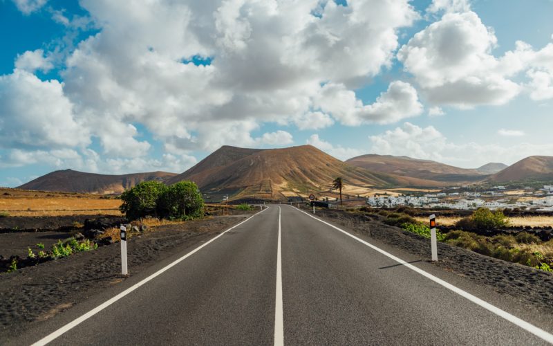 looking down a grey asphalt road towards brown mountains with a small town visible in the distance on the right and a few small trees along the road with lots of fluffy clouds overhead. best areas in lanzarote - where to stay.