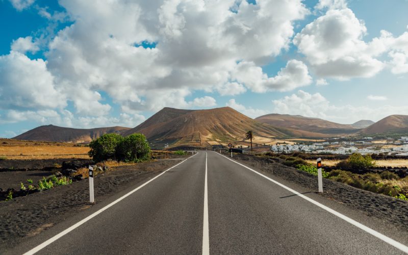 lookign down a grey asphalt road towards brown mountains with a small town visible in the distance on the right and a few small trees along the road with lots of fluffy clouds overhead. best areas in lanzarote - where to stay. 