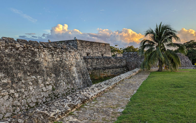 grey stone walls of the exterior of a historic spanish fort in bacalar mexico with a single palm tree onthe lawn outside and the sky turning golden just before sunset