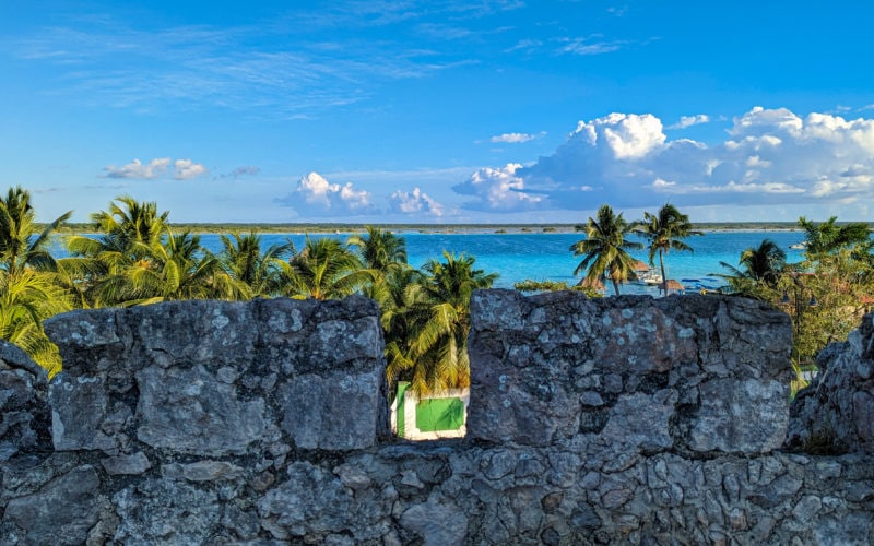 view from san felipe fort in bacalar mexico looking out between the grey stone crenellations at the bright blue lake behind a dense patch of green palm trees