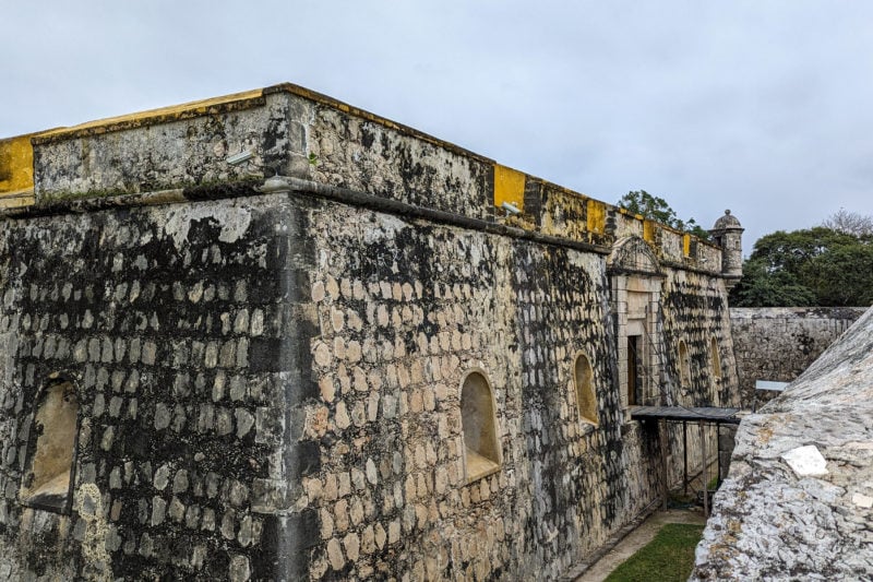 corner of the exterior stone wall of san jose fort in campeche with an empty moat filled with grass around the outside and a wooden drawbridge over the moat leading to the main door.