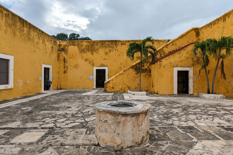 inside of San Migue Fort in Campeche, a grey flagstone floor with a small stone well in the middle surrounded by walls painted a dark yellow with grey sky overhead