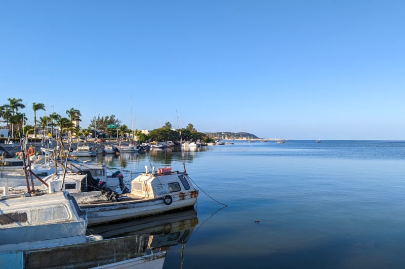 view of the sea with a very flat calm surface and vivid blue water on a sunny day with blue sky above and several small white fishing boats moored near the sea wall to the left.