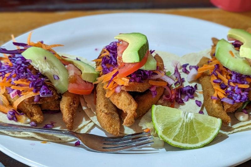 close up of 3 tacos on a white plate with battered fried strips of fish in the tacos and red cabbage and slices of tomato and avocado on top and a lime wedge next to the tacos