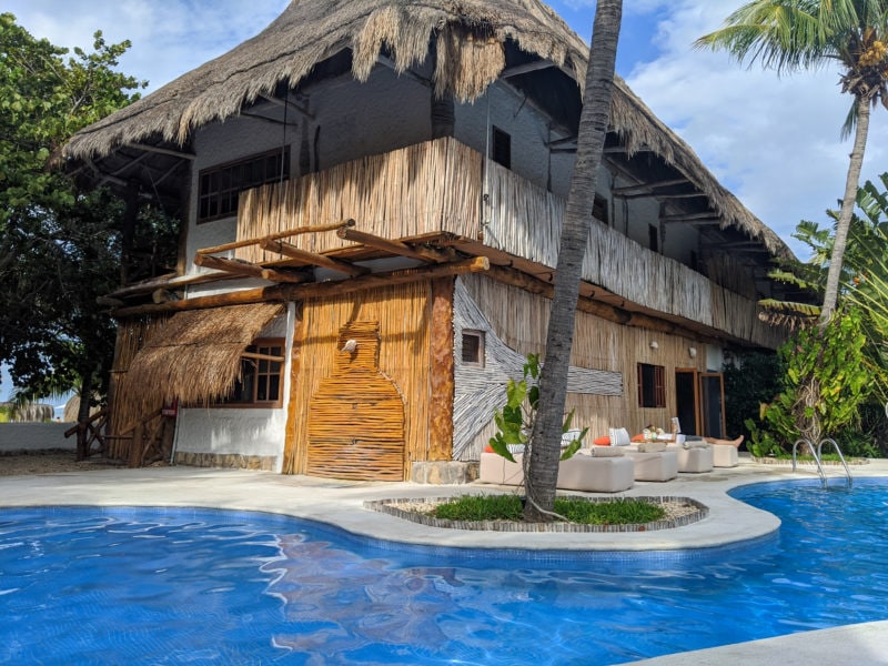 curved blue hotel pool outside with palm trees dotted around and a laerge two story whitewashed building on the far side with grass frond roof an a long balcony with wooden fence.  