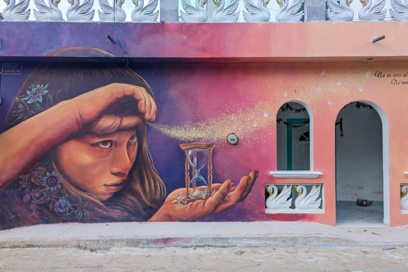 large street art mural of a mexican girl with long dark hair holding an hourglass and sprinkling sand onto the wind against a pink and purple background on a wall next to a sand road. things to do in holbox mexico