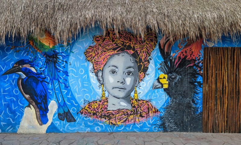 street art mural of a girl in an orange headress and orange shawl with long earings on a blue background surrounded by birds. the mural is on the side of a building with a thatched grass roof.