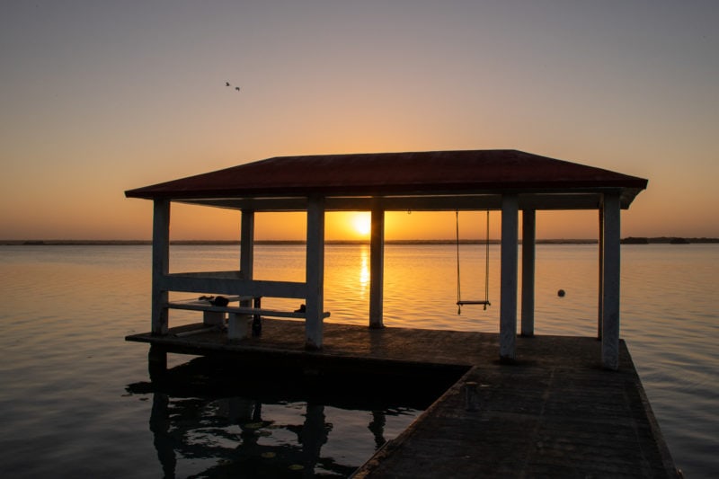 silhouette of a wooden jetty with a covered structure and a swing at the end in a very flat calm lake at sunrise with the golden sun just above the horizon reflected in the lake. things to do in Bacalar.